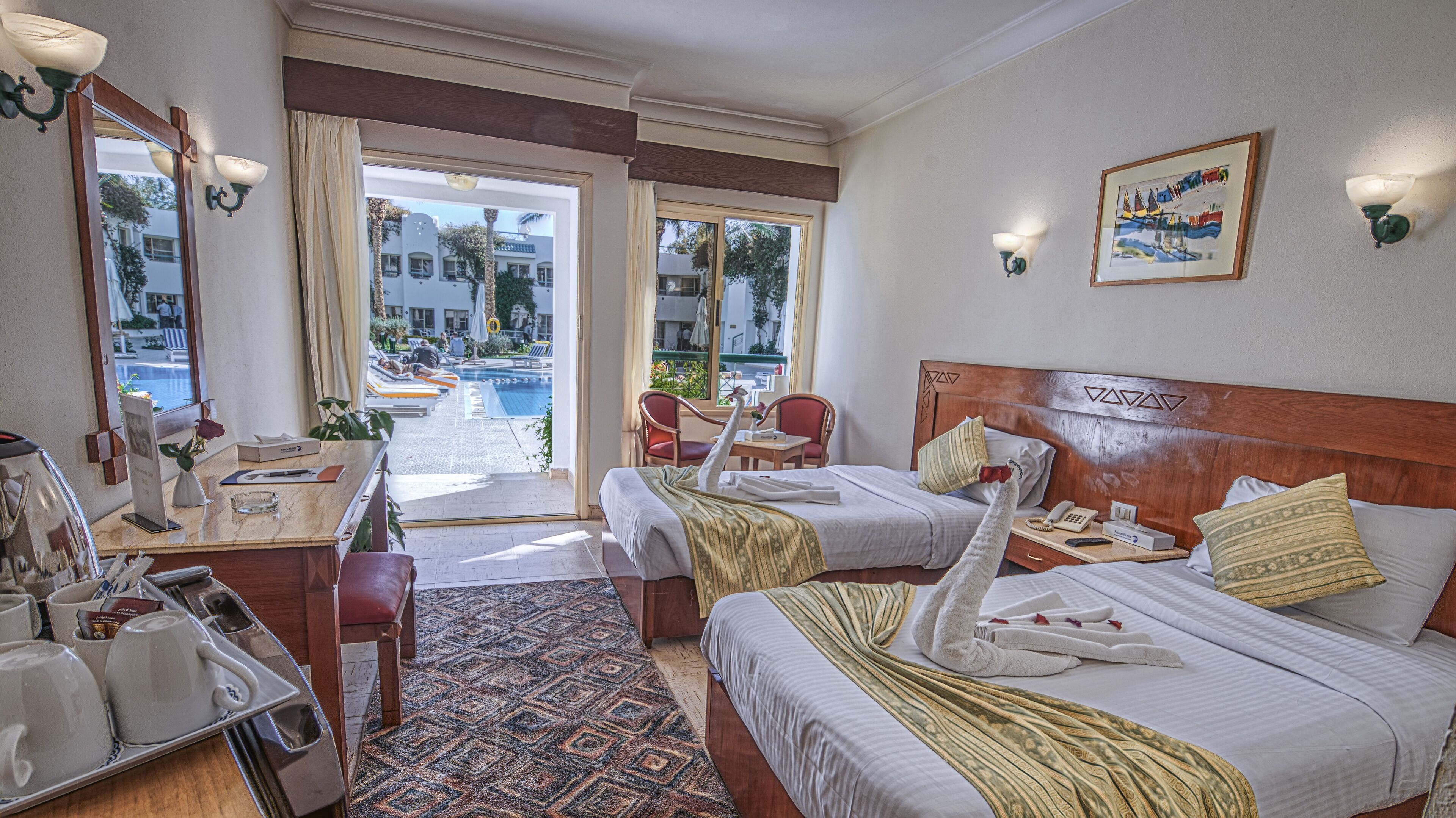 A hotel room with two beds, a desk with a chair, a small table, a mirror, and a view of an outdoor pool area.