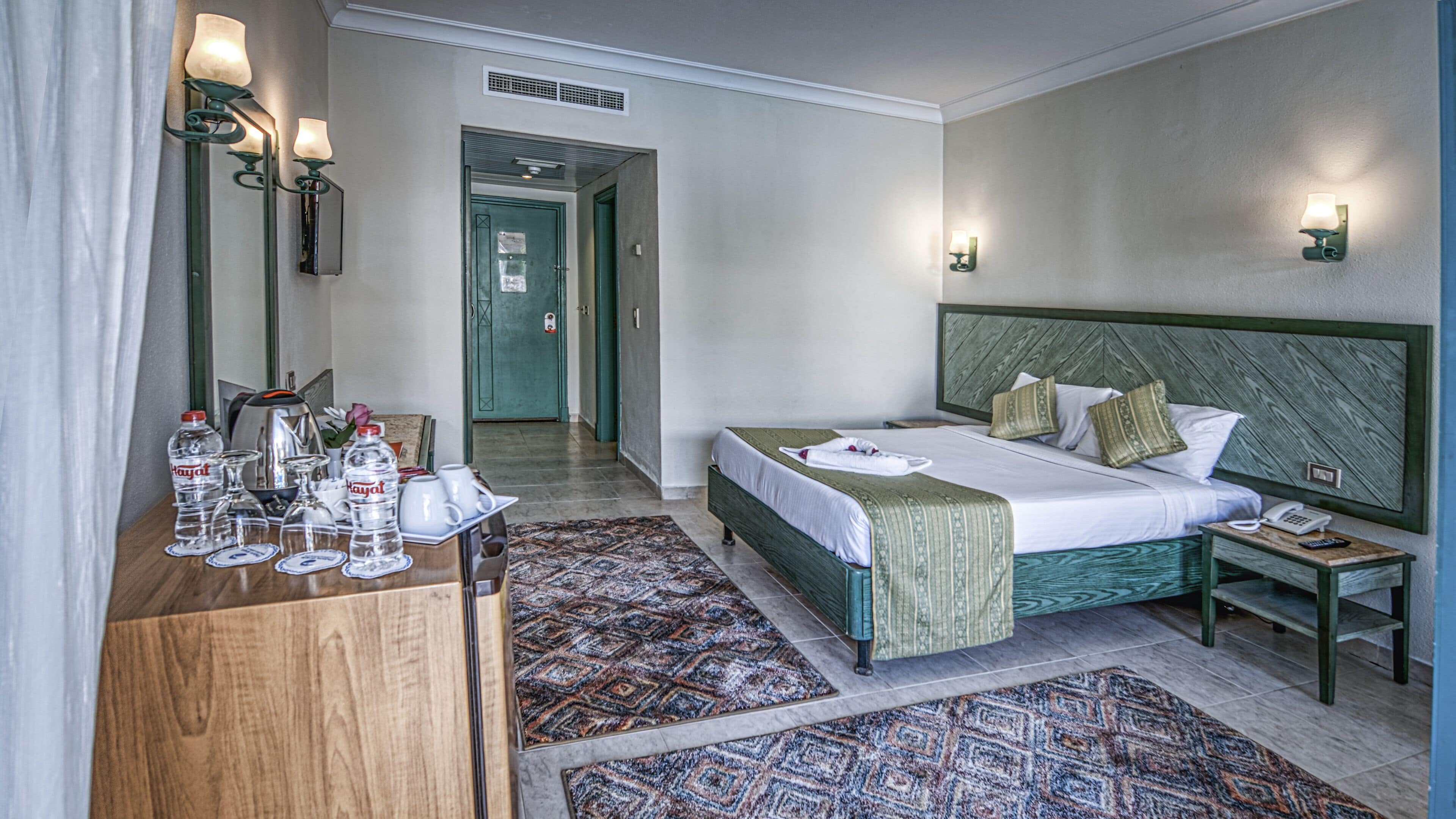 A hotel room with a bed, bedside table, lamp, and a desk with a mirror and toiletries.