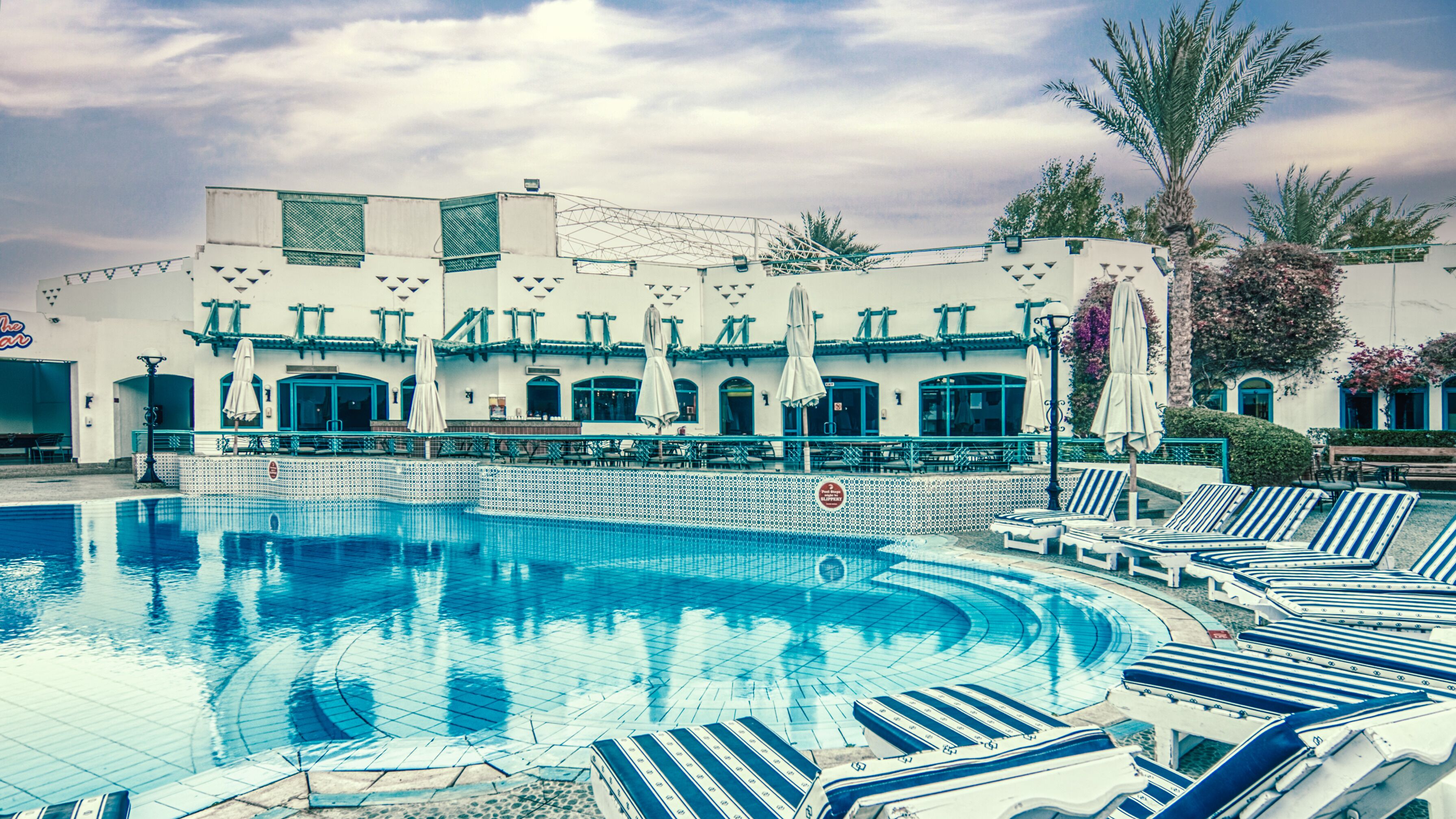 Ein Swimmingpool mit Liegestühlen und Sonnenschirmen, umgeben von einem Gebäude mit einer Kachelfassade und Außenbereich.