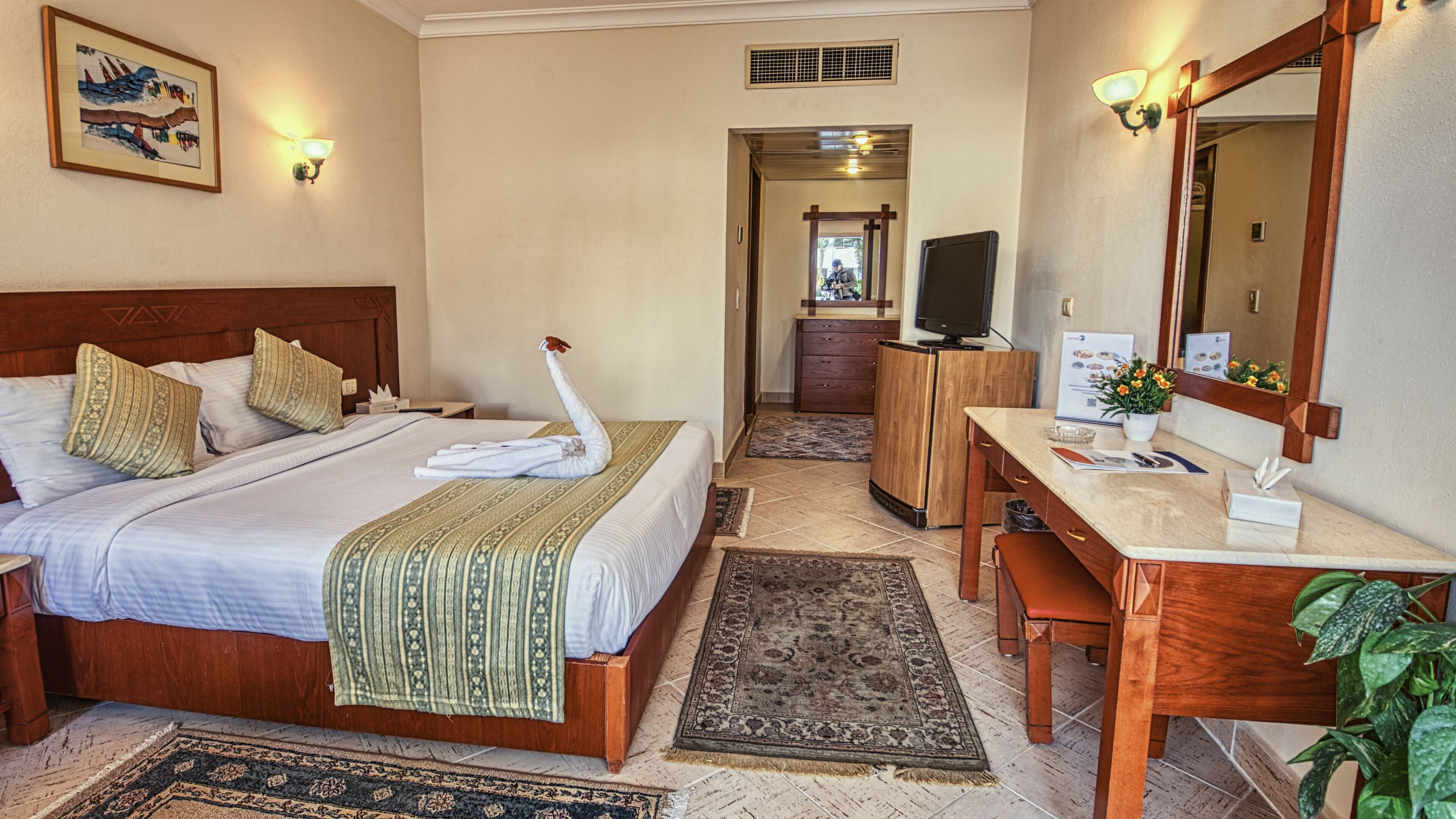 A hotel room with a bed, a desk, a mirror, a television, and a bathroom visible in the background.