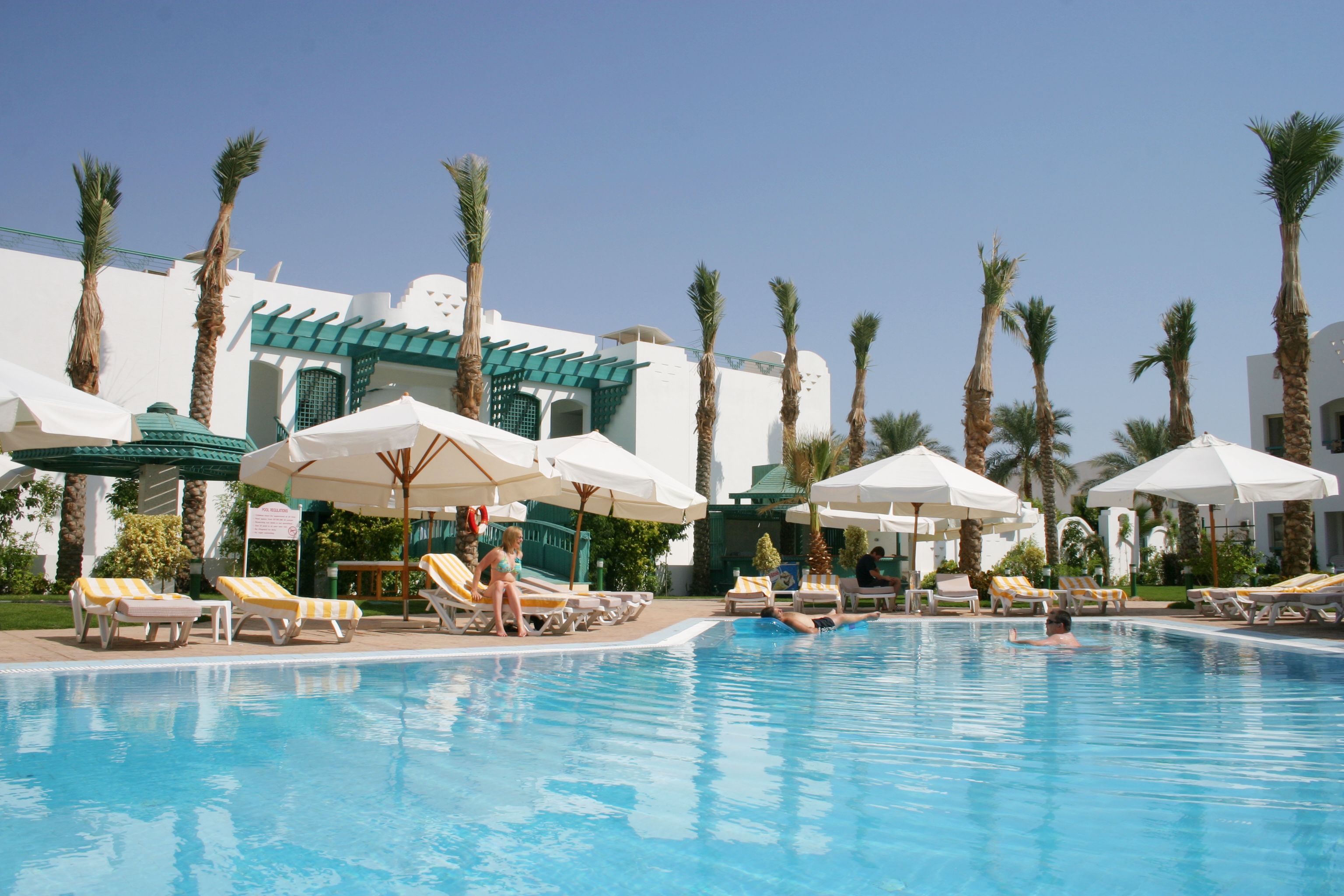 A swimming pool with lounge chairs and umbrellas, surrounded by palm trees and a building with a white facade.