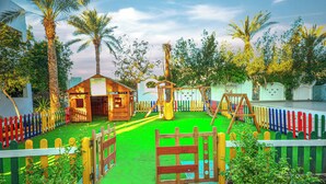 A playground with a wooden playhouse, swings, and a slide, surrounded by a colorful fence and palm trees. - Falcon Hills Hotel (Sharm El Sheikh)