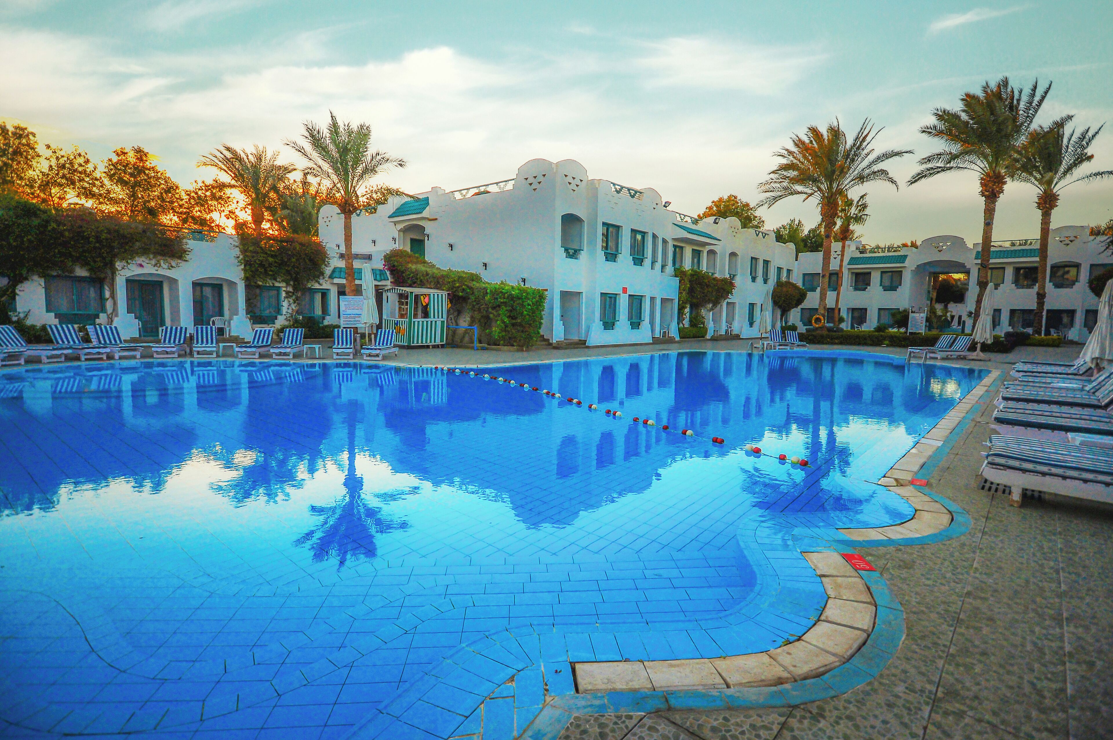 A swimming pool with lounge chairs and palm trees, surrounded by buildings with a Mediterranean architectural style.