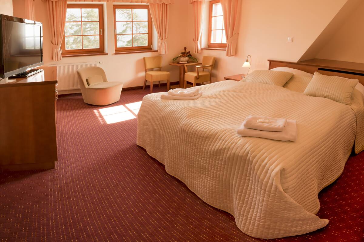 A hotel room with a large bed, a desk with a TV, two chairs, a small table, and windows with curtains.