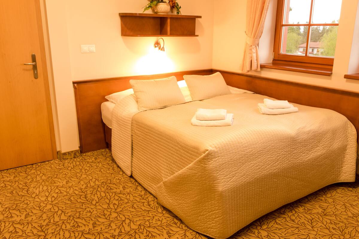A neatly made bed with a headboard, a bedside table with a lamp, and a window with curtains.
