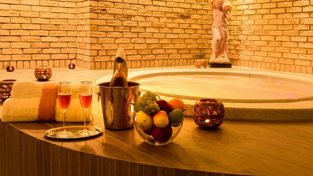 Zona de spa con pared de ladrillo, una estatua, bañera de hidromasaje, frutero y dos vasos de bebida roja sobre una bandeja.
