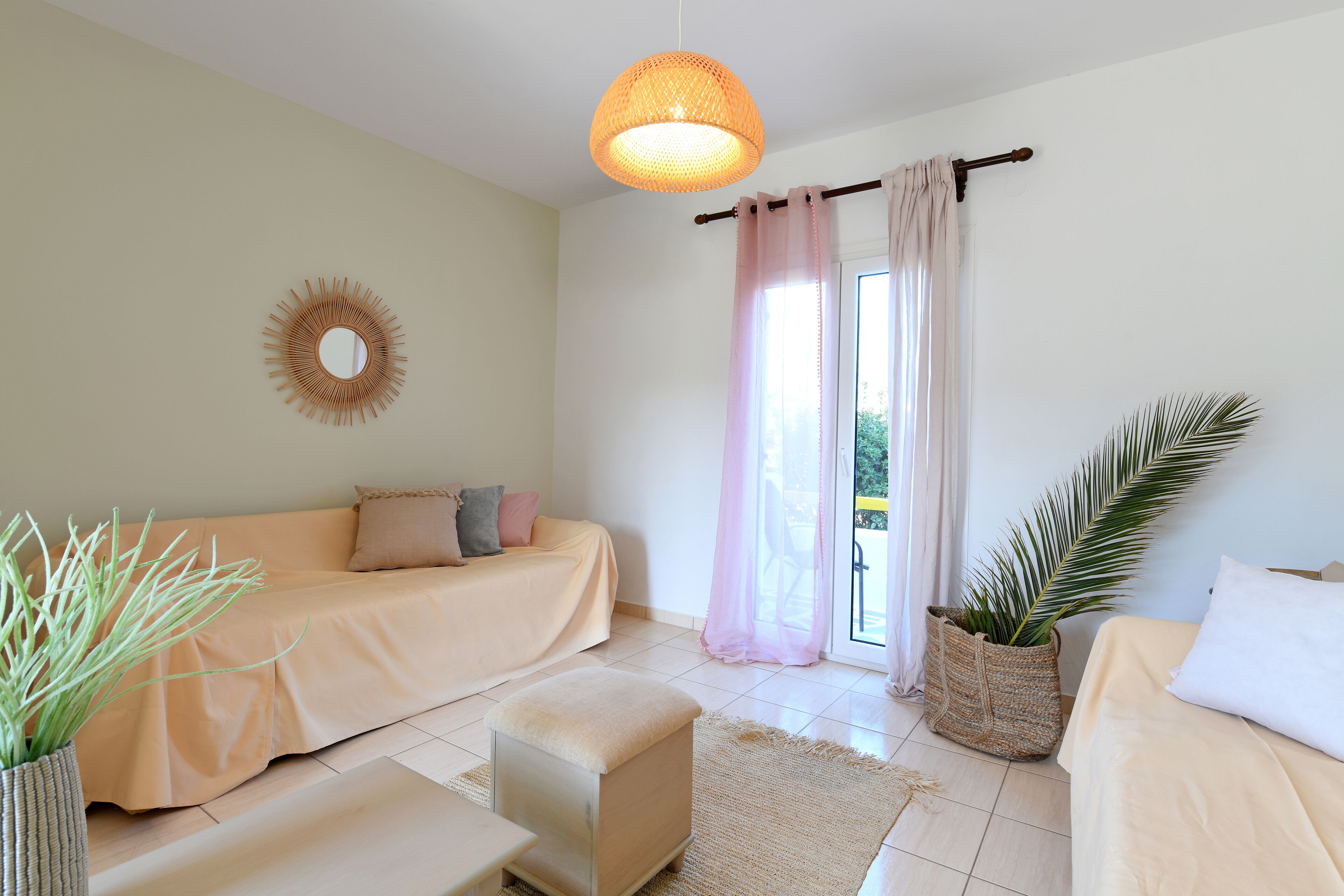 Un dormitorio moderno con un sofá beige, un pequeño otoman, una lámpara de pie trenzada y una planta de palma en maceta.
