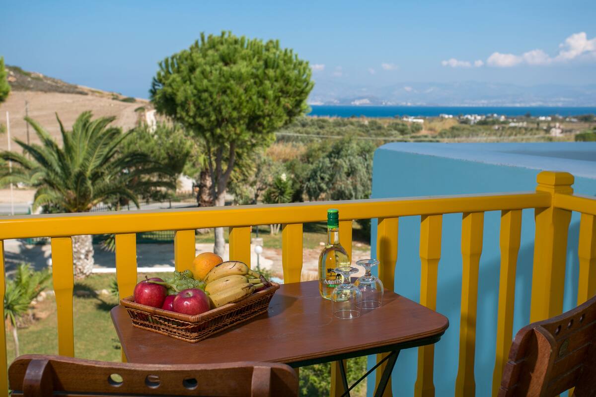 A balcony with a table set with fruit and wine glasses, overlooking a scenic landscape with trees and a body of water.