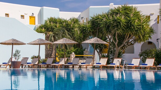 A pool area with lounge chairs and umbrellas, surrounded by palm trees and a building with white and yellow walls.