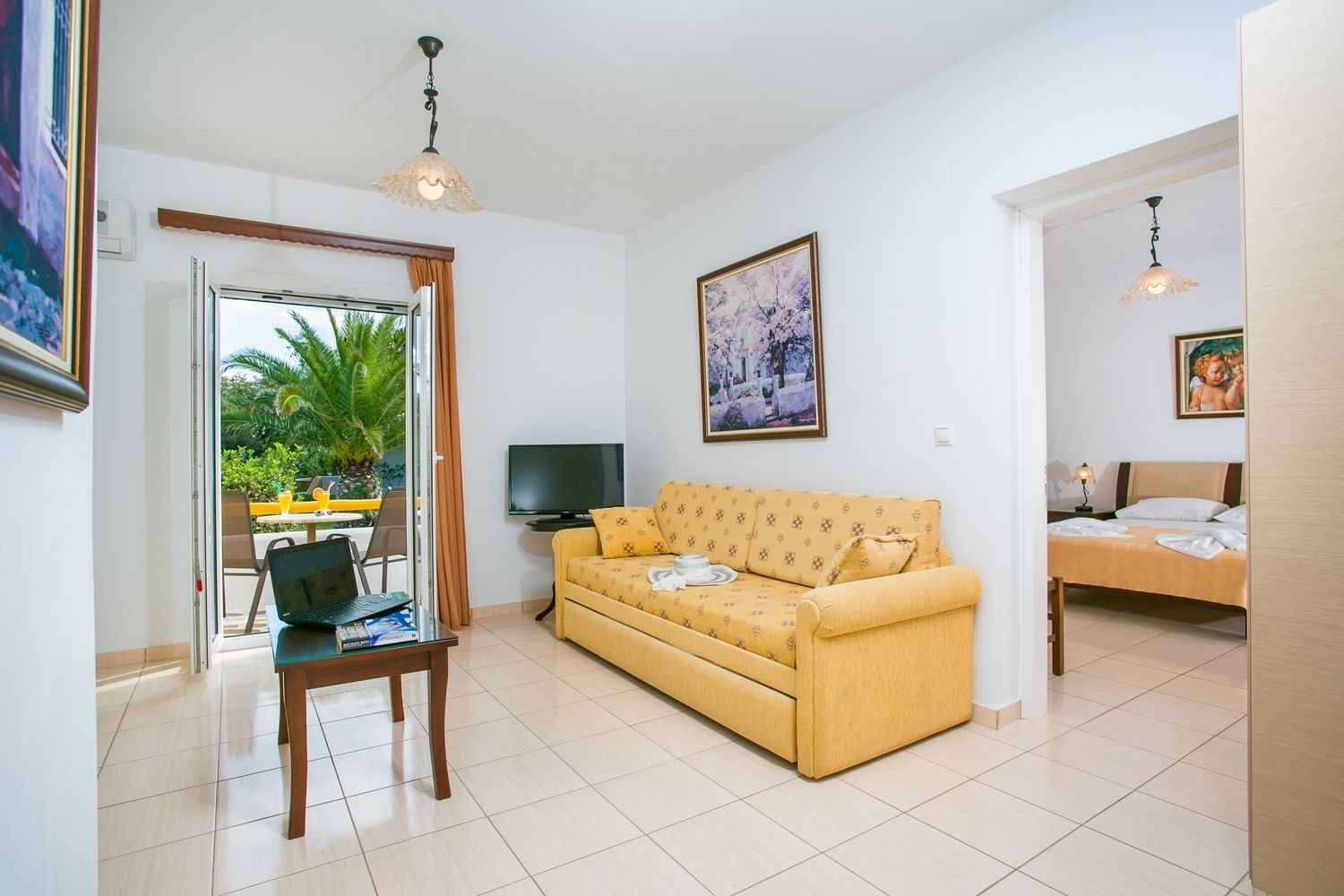 Una sala de estar con un sofá amarillo, un televisor, una mesa de comedor con sillas y un balcón con vistas a palmeras.