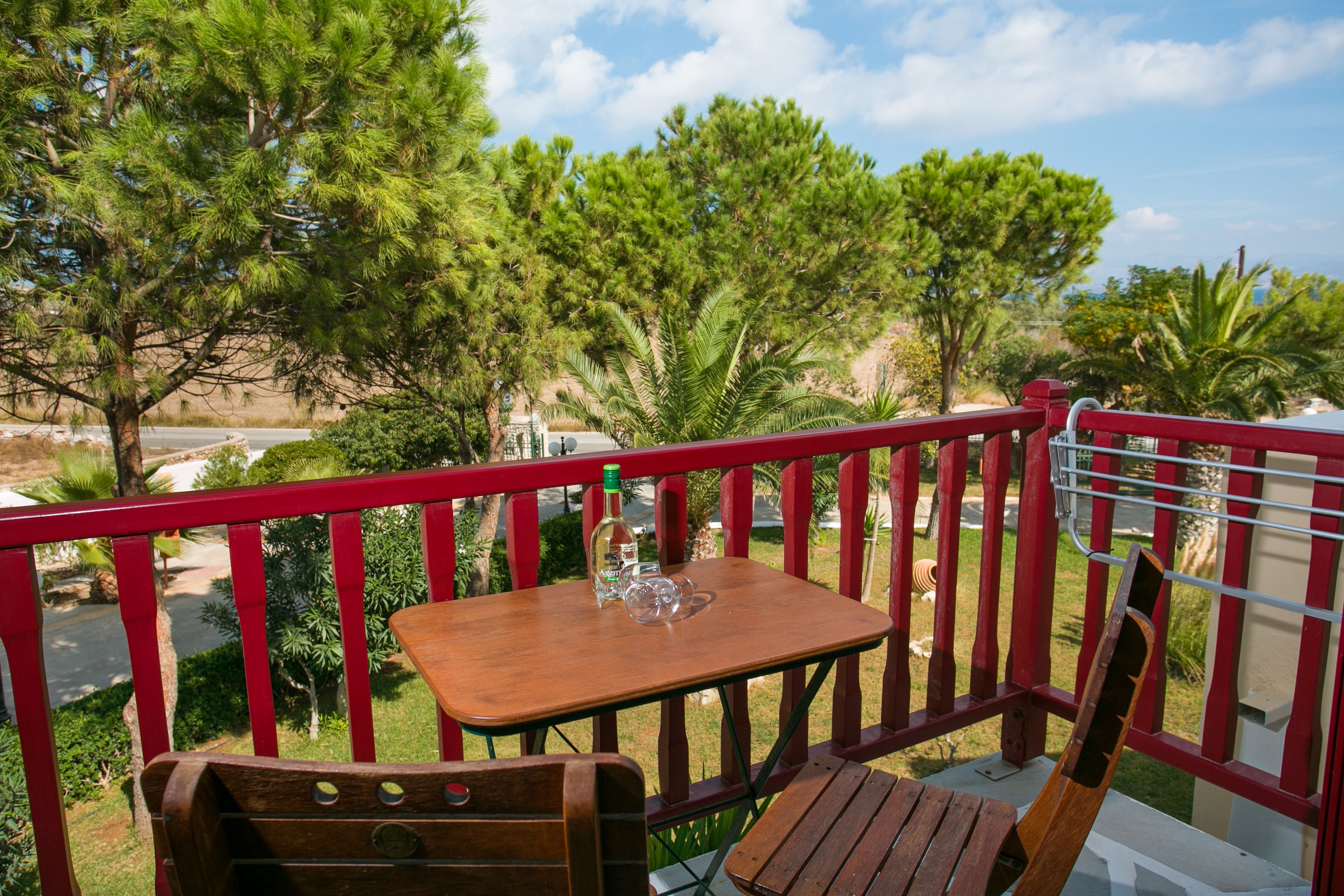 A wooden table with a bottle and glass on a balcony with red railings, overlooking a road and greenery.