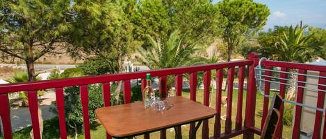 A wooden table with a bottle and glass on a balcony with red railings, overlooking a road and greenery.