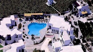 An aerial view of a resort with a central swimming pool, surrounded by white buildings with arched windows and a few palm trees.