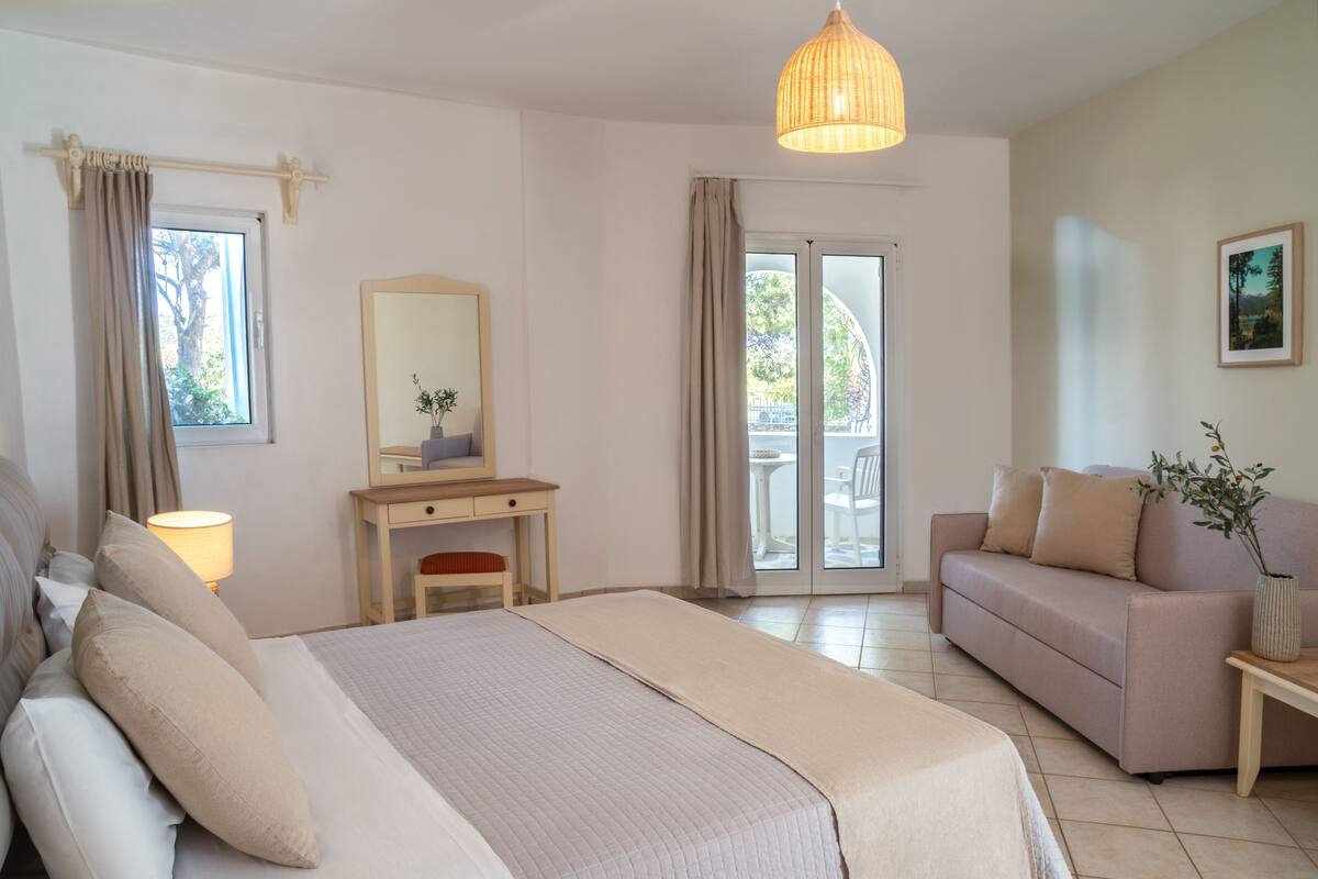 A hotel room with a bed, a desk, a sofa, a large window, and a balcony.