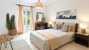 A bedroom with a large bed, bedside tables, a desk, a chair, a hanging light fixture, and a view of the outdoors.