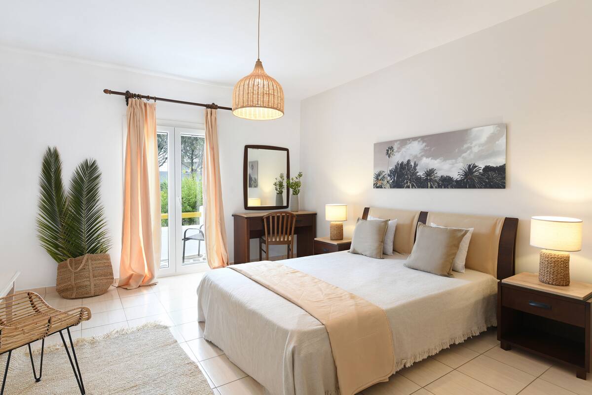 A bedroom with a large bed, bedside tables, a desk, a chair, a hanging light fixture, and a view of the outdoors.