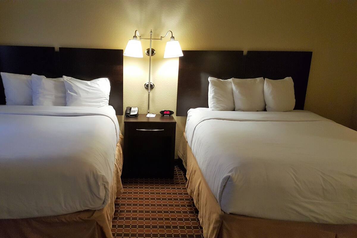 Una habitación de hotel con dos camas, una mesita de noche con teléfono y reloj, y una lámpara fijada a la pared.