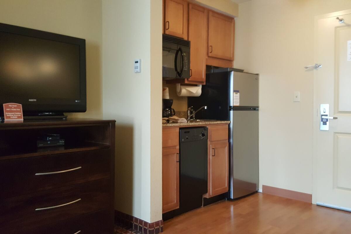 Una cocina compacta con armarios de madera, microondas integrado, refrigerador y lavavajillas. Un televisor de pantalla plana está montado en un mueble de madera.