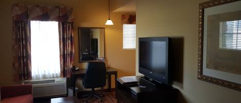 Habitación de hotel con televisión de pantalla plana, escritorio, silla, sofá y una ventana grande con persianas.