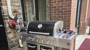 Una parrilla de gas portátil con dos hornos, un termómetro y un quemador lateral, ubicada frente a un edificio de ladrillo con una ventana y un hidrante rojo a la vista.