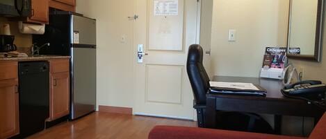 A compact kitchen with a refrigerator, dishwasher, and microwave. There's a door with an emergency evacuation plan posted. A desk with a chair and a phone is visible, along with a magazine rack.