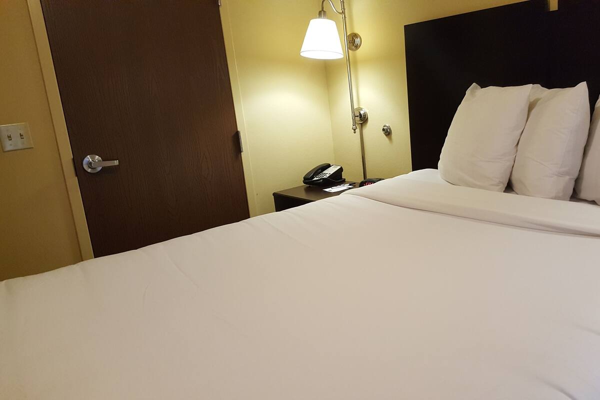 Una cama bien hecha con sábanas y almohadas blancas, un teléfono en la mesita de noche y una lámpara fijada a la pared.