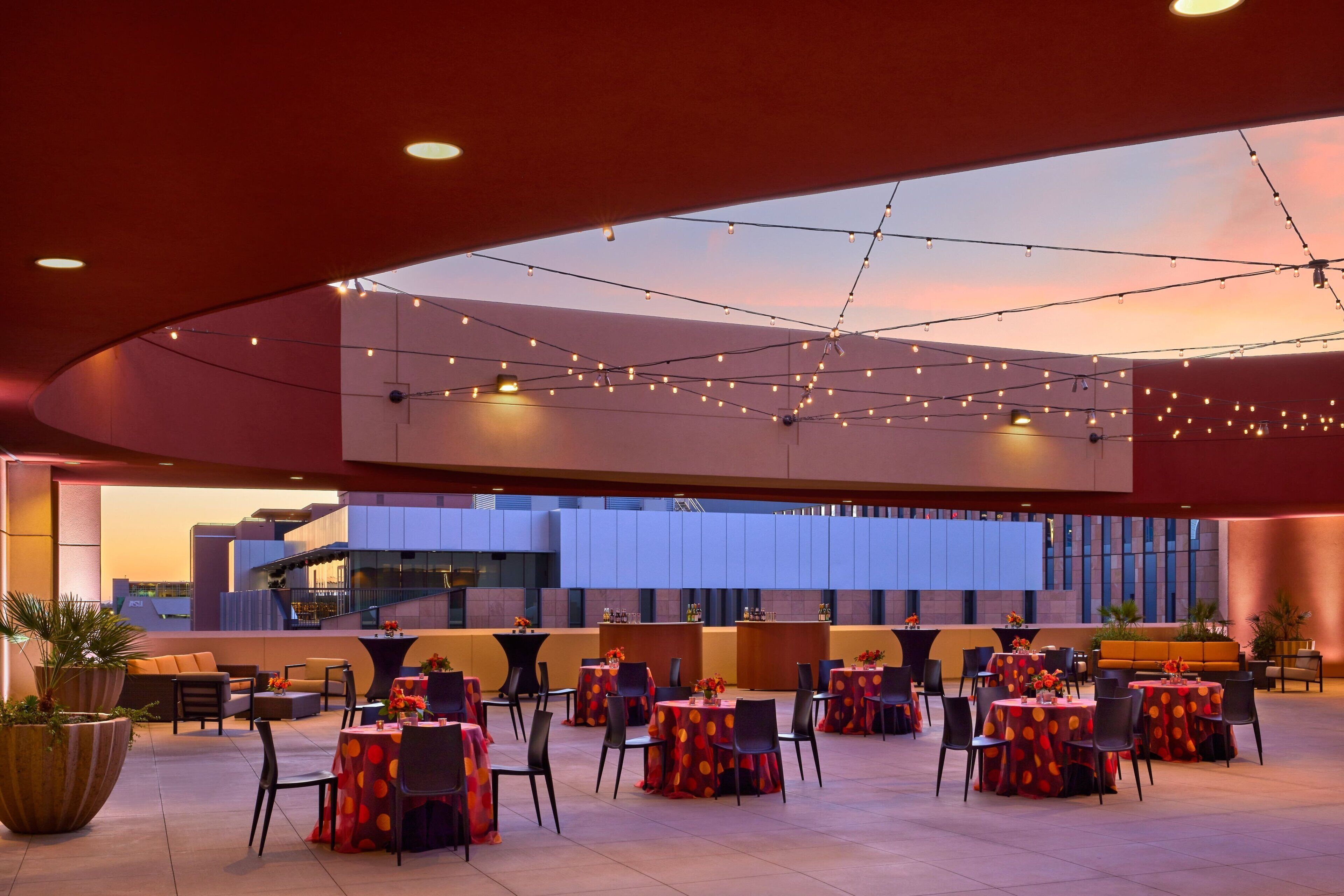 A spacious outdoor dining area with multiple tables and chairs, string lights, and a modern building in the background.