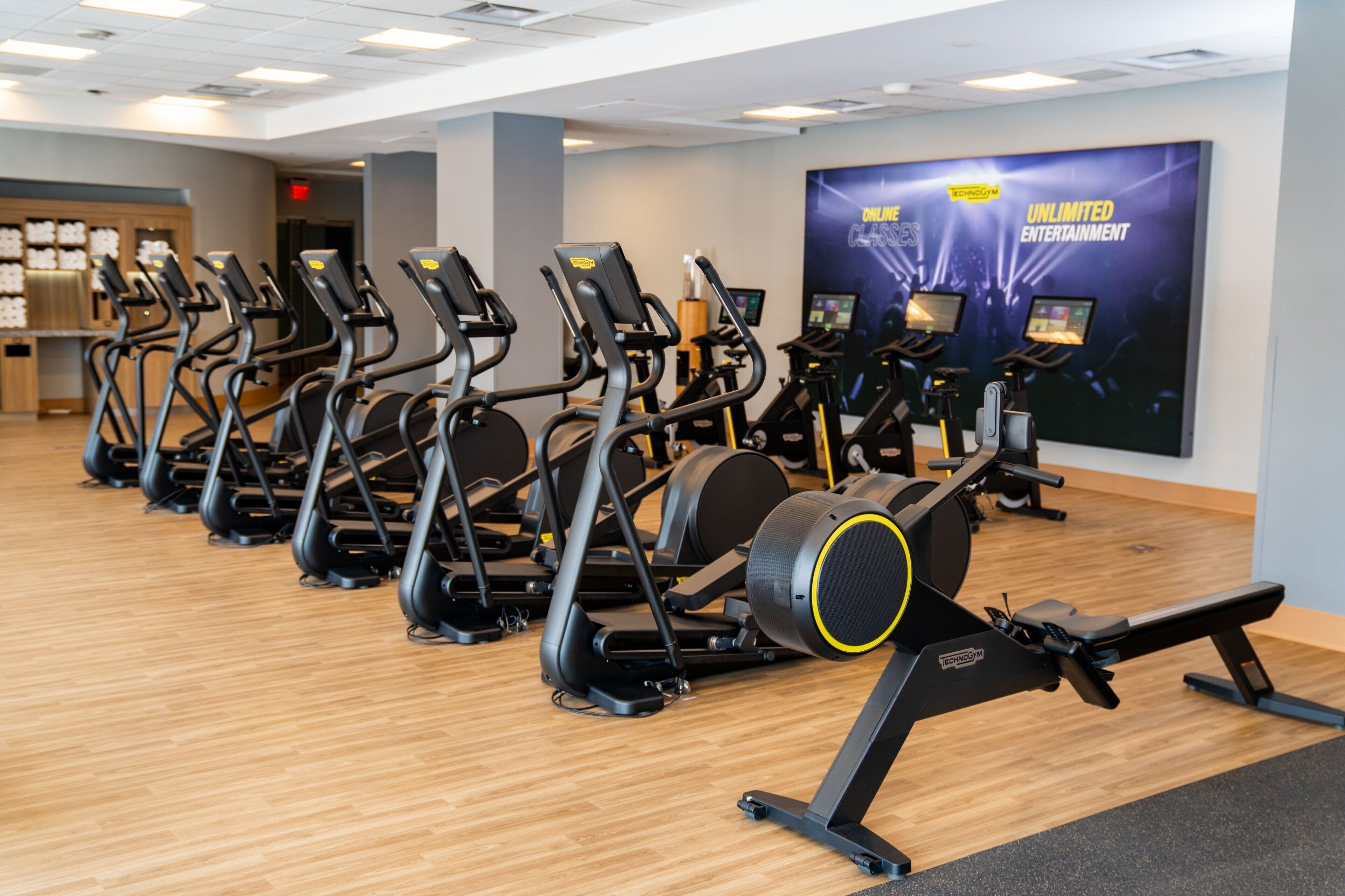 A gym with multiple treadmills and elliptical machines, wooden flooring, and a large poster advertising unlimited entertainment.