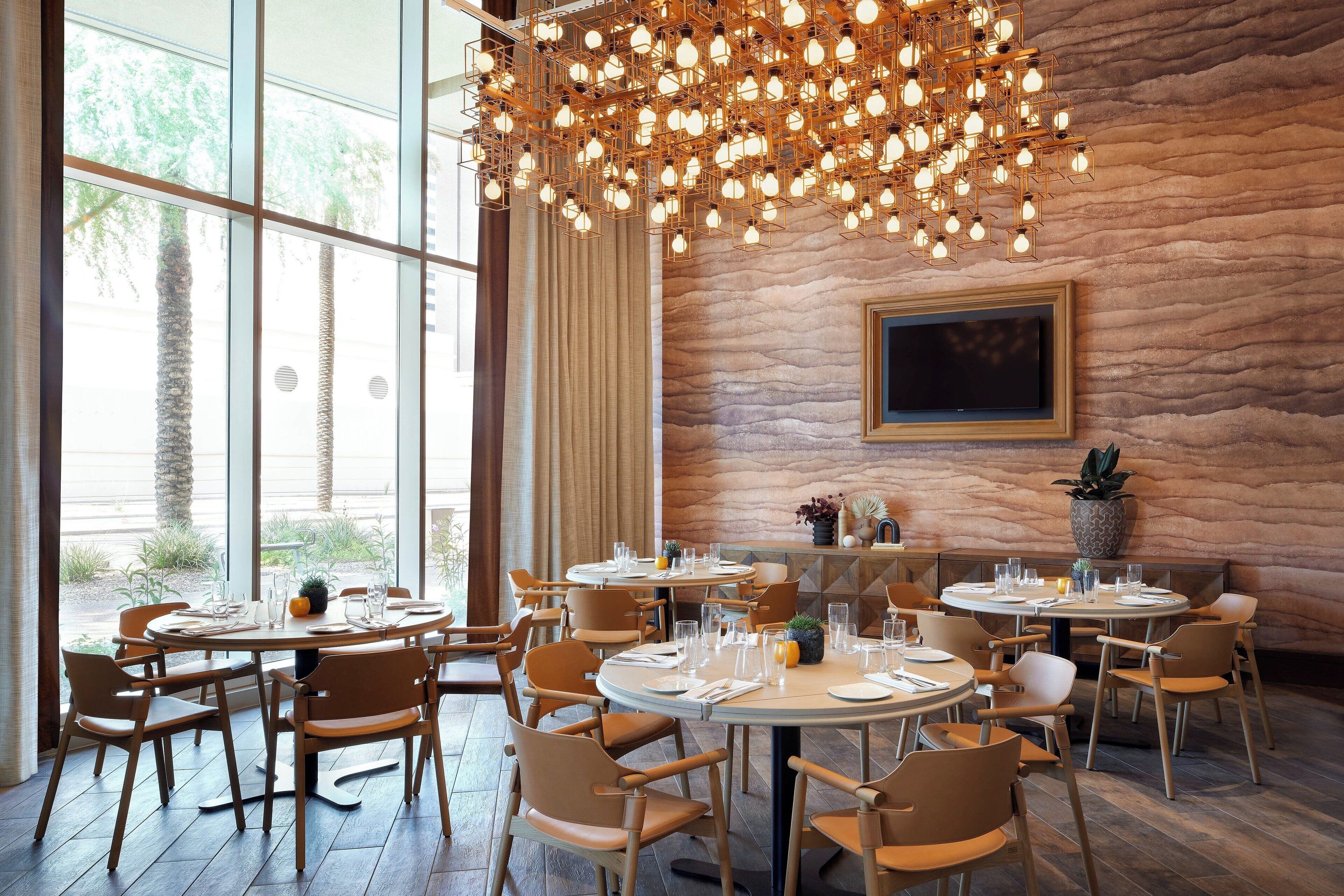 A restaurant with round tables and chairs, a large chandelier, and a stone wall.