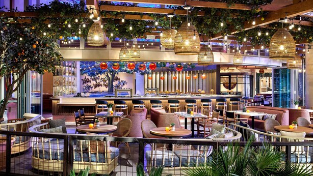 Un restaurante interior con varias áreas de comedor, lámparas de mimbre colgantes y plantas de interior.