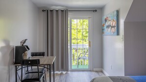 Une pièce avec une porte-fenêtre coulissante donnant sur un balcon, une table avec des chaises, un bureau avec une lampe et un tableau au mur.