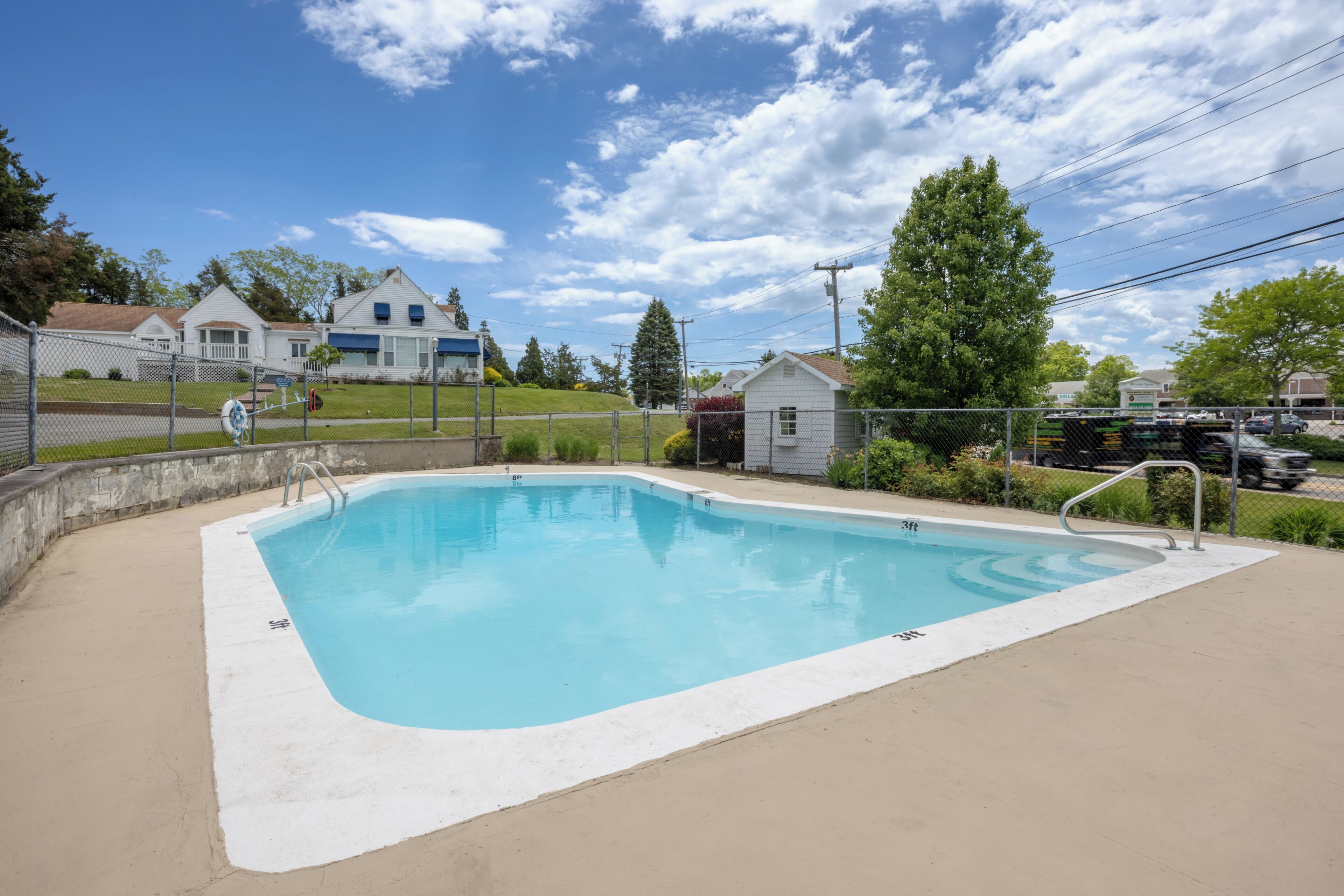 En rektangulær swimmingpool med en betonbelagt terrasse, omgivet af et hegn af pigtråd, med huse og et stort træ i baggrunden.