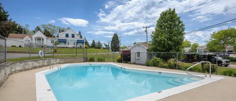 En rektangulær swimmingpool med en betonbelagt terrasse, omgivet af et hegn af pigtråd, med huse og et stort træ i baggrunden.