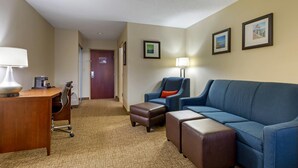 A hotel room with a desk, chair, blue sofa, ottoman, and a hallway leading to a door.