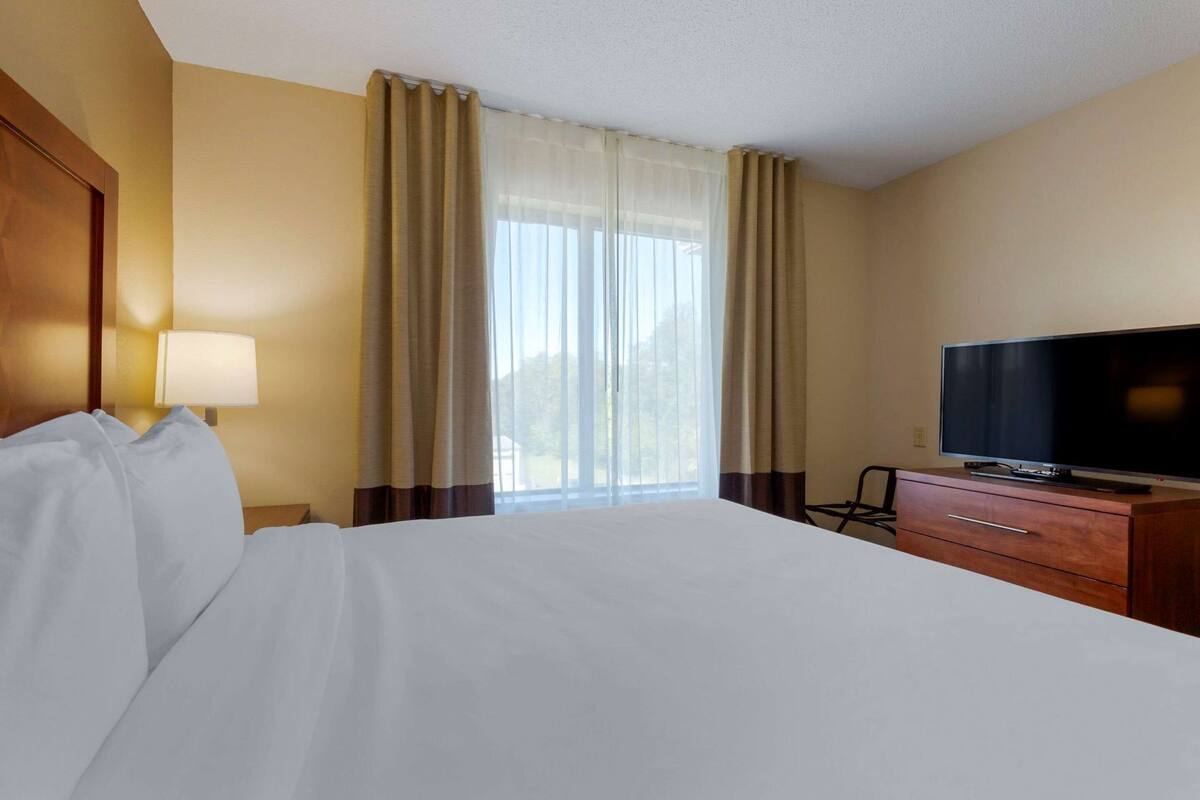 Ein Hotelzimmer mit einem Bett, einem Fernseher auf einer Holzkommode, einer Lampe und einem Fenster mit Vorhängen.