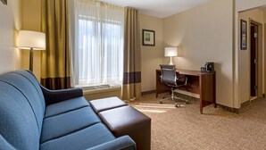 A hotel room with a blue sofa, a desk with a chair, a lamp, and a picture on the wall.