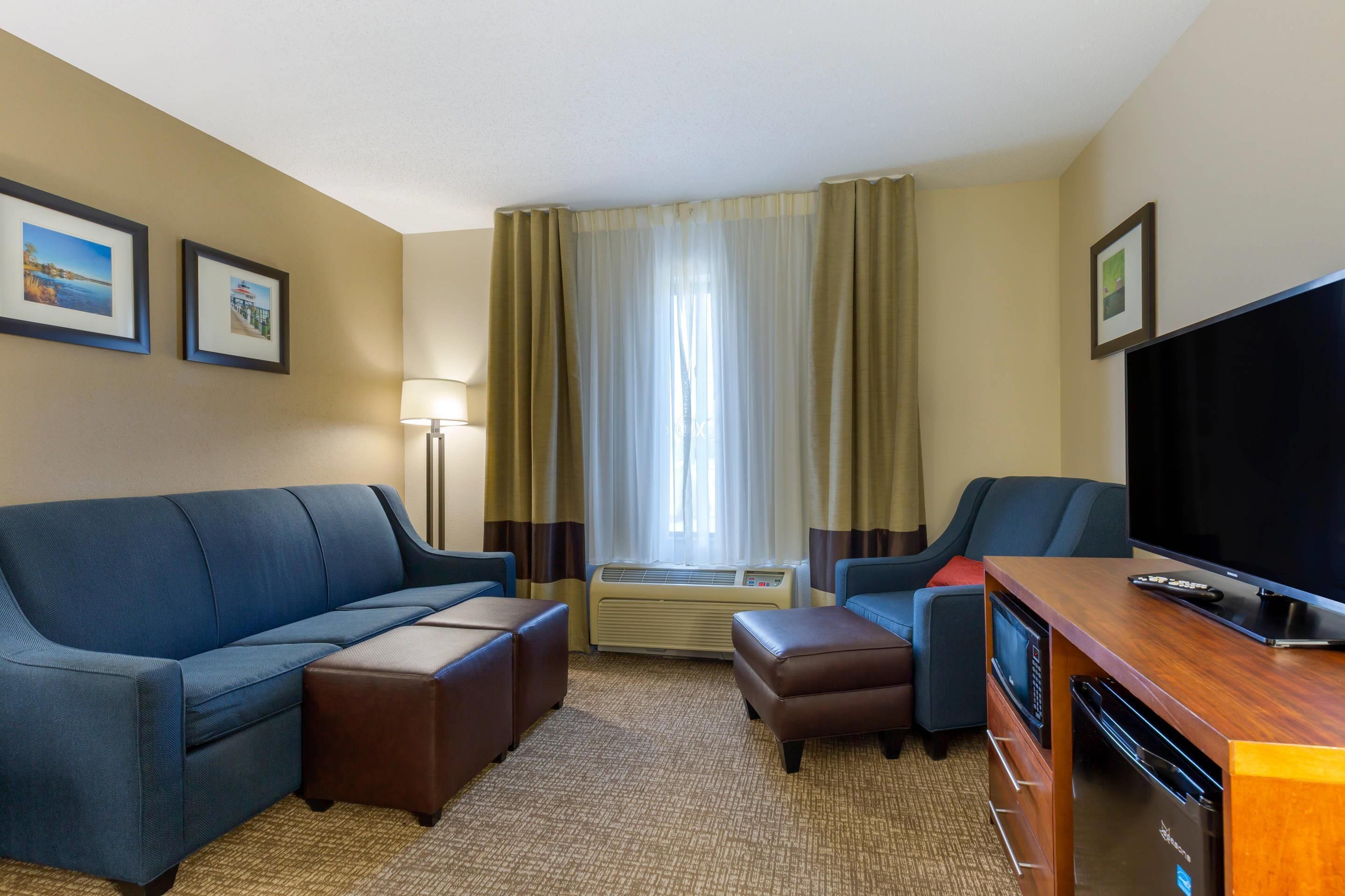 A hotel room with a blue sofa, a brown ottoman, a wooden TV stand, and a flat-screen TV.