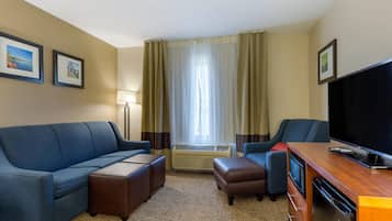 A hotel room with a blue sofa, a brown ottoman, a wooden TV stand, and a flat-screen TV.