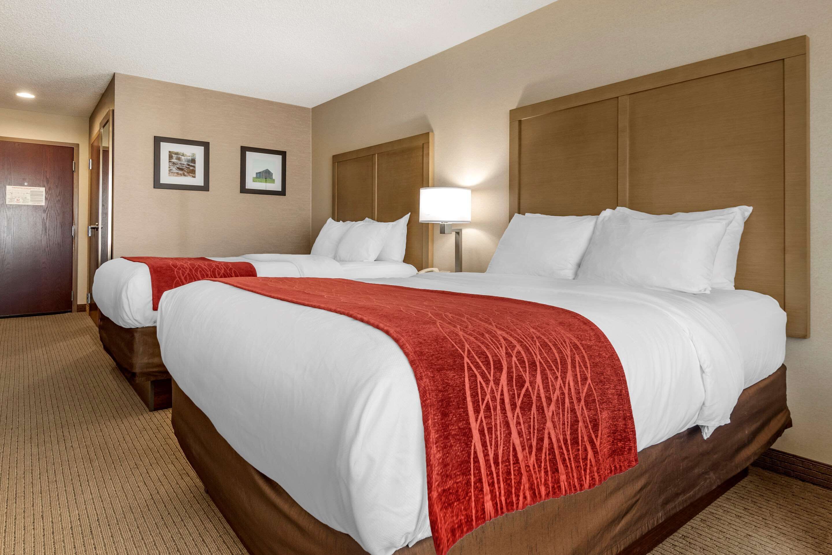 A hotel room with two beds, a wooden headboard, a red throw blanket, and a wall-mounted lamp.