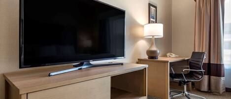 A hotel room with a large flat-screen TV, a desk with a lamp, a chair, and a telephone.