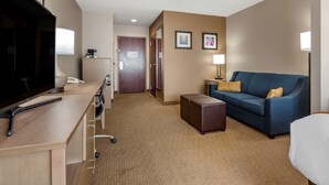 Ett hotellrum med en platt-TV, ett skrivbord med telefon och kaffebryggare, en blå soffa med kuddar och en liten pall.