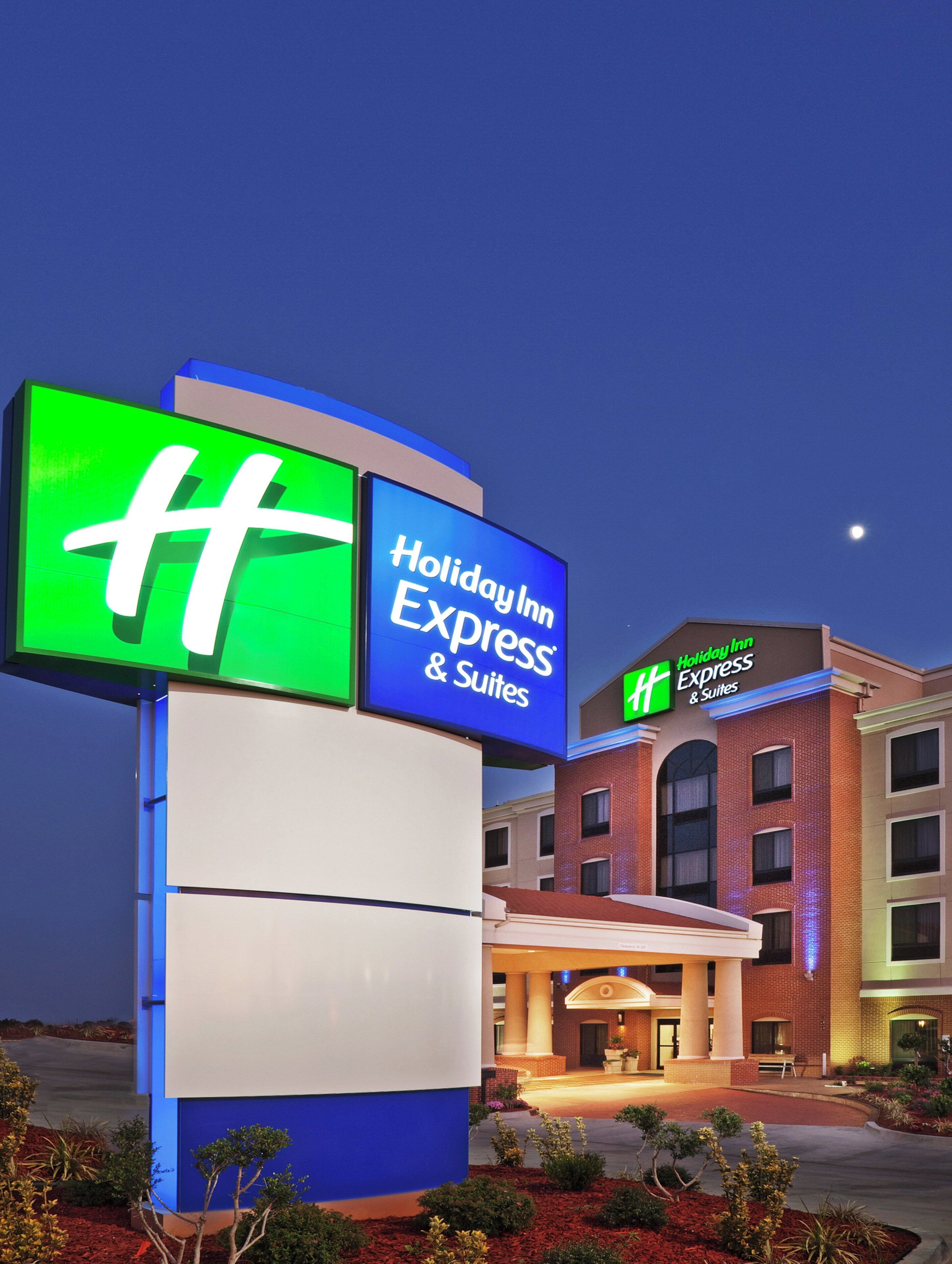 Placa do Holiday Inn Express & Suites com o logo 'H' em destaque, entrada de um edifício e céu noturno claro.