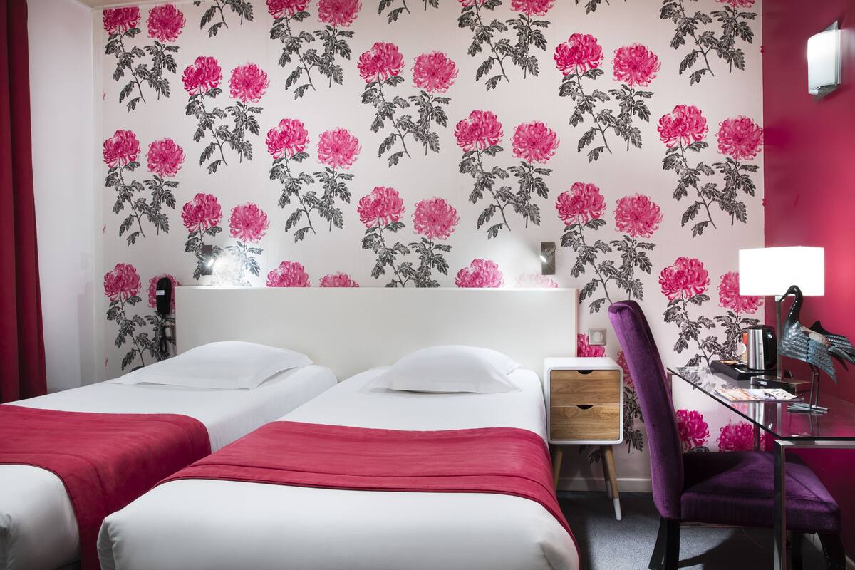 Une chambre d’hôtel avec deux lits, un bureau avec une chaise, une table de chevet et un mur recouvert d’un papier peint à motifs floraux.