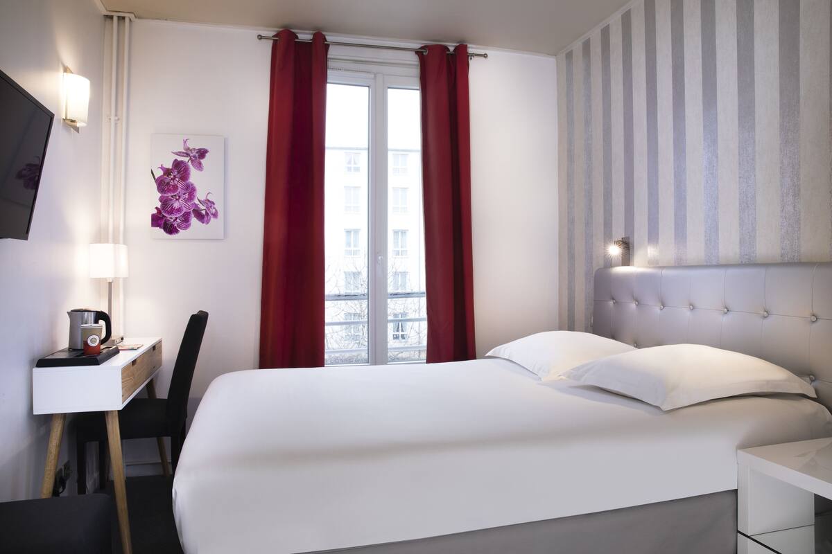 Une chambre d’hôtel comprenant un lit, un bureau avec une chaise, une lampe, un tableau représentant des fleurs et une fenêtre avec des rideaux.