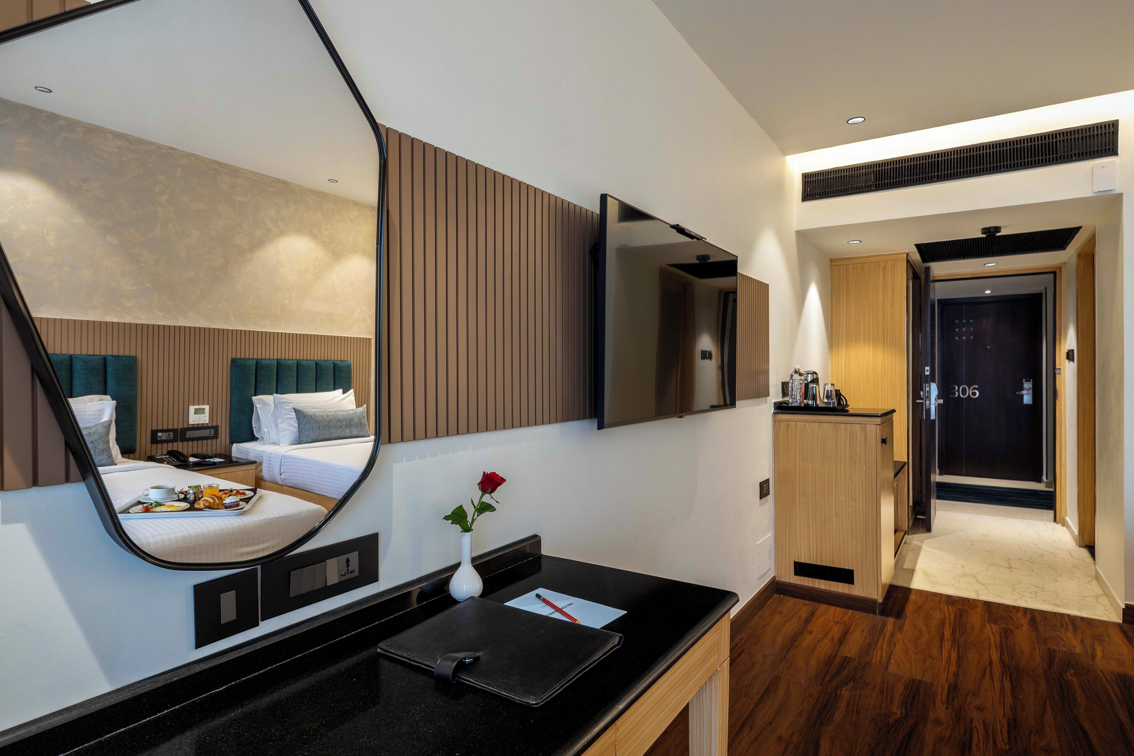 Una habitación de hotel con un espejo grande, una mesita de noche con una bandeja de desayuno, un jarrón con una rosa roja y una puerta de madera con el número 306.
