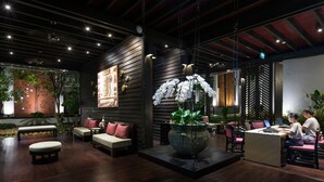 Un interior moderno con piso de madera, una zona de estar central con cojines, una jardinera grande con flores blancas y un escritorio con dos personas trabajando.