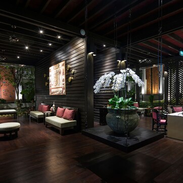 A modern interior with a wooden floor, a central seating area with cushions, a large planter with white flowers, and a desk with two people working.