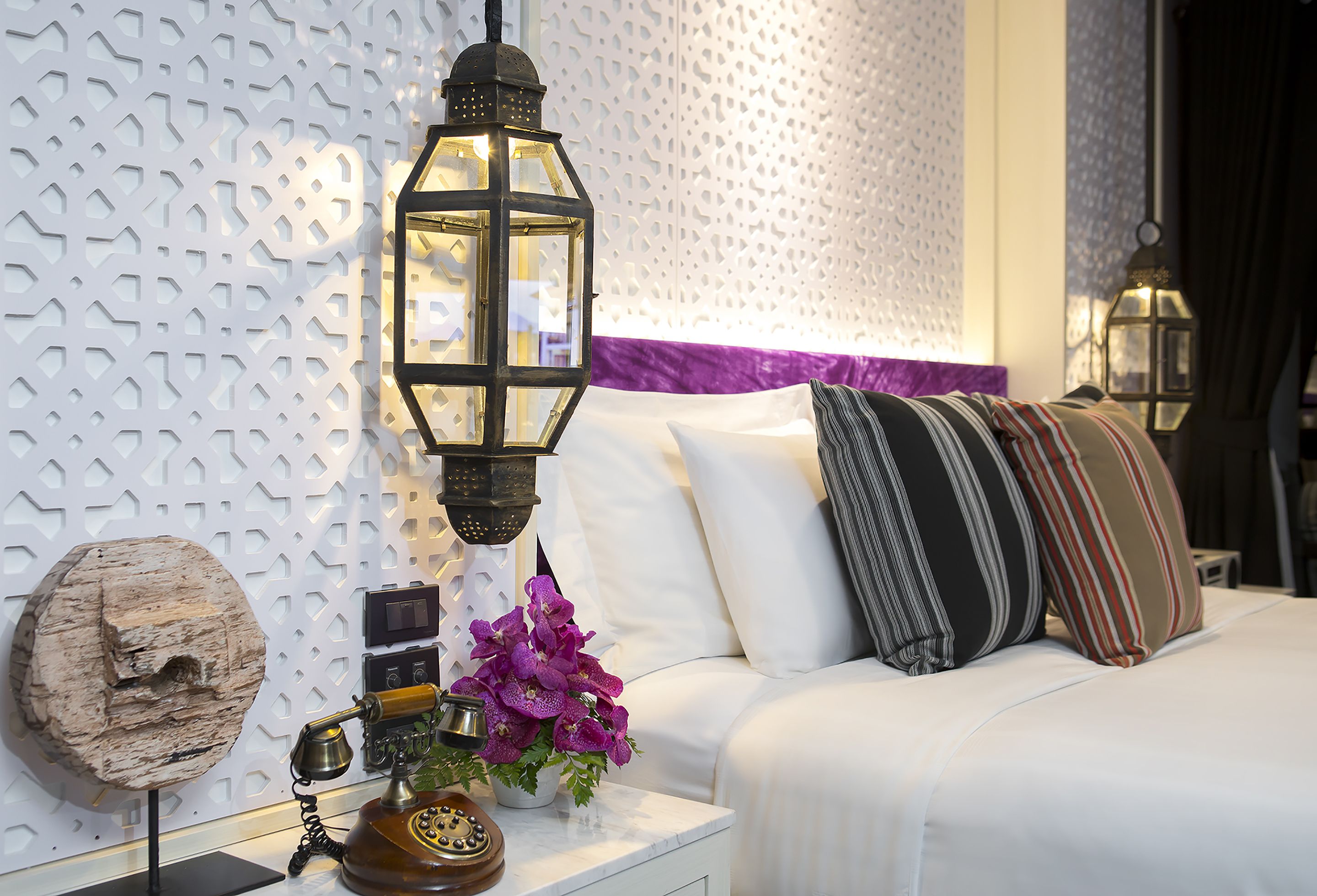 A neatly arranged bedroom with a white bed, decorative pillows, a wall-mounted light fixture, and a vintage telephone on a bedside table.