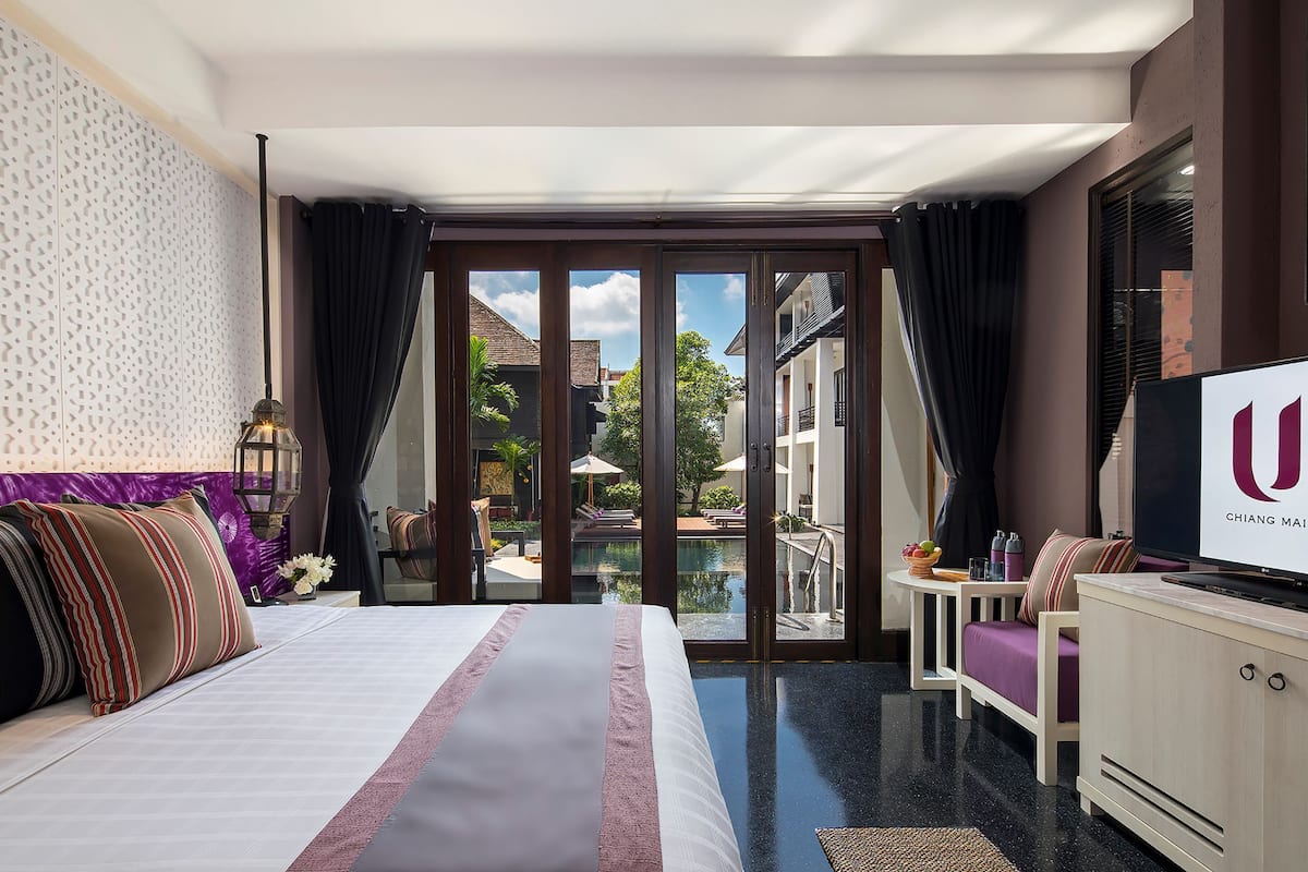 A hotel room with a bed, a TV, a desk, and a view of a pool and buildings outside.