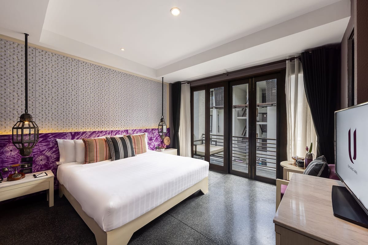 A hotel room with a large bed, a desk with a computer, a TV, a balcony with a view, and a patterned wall.
